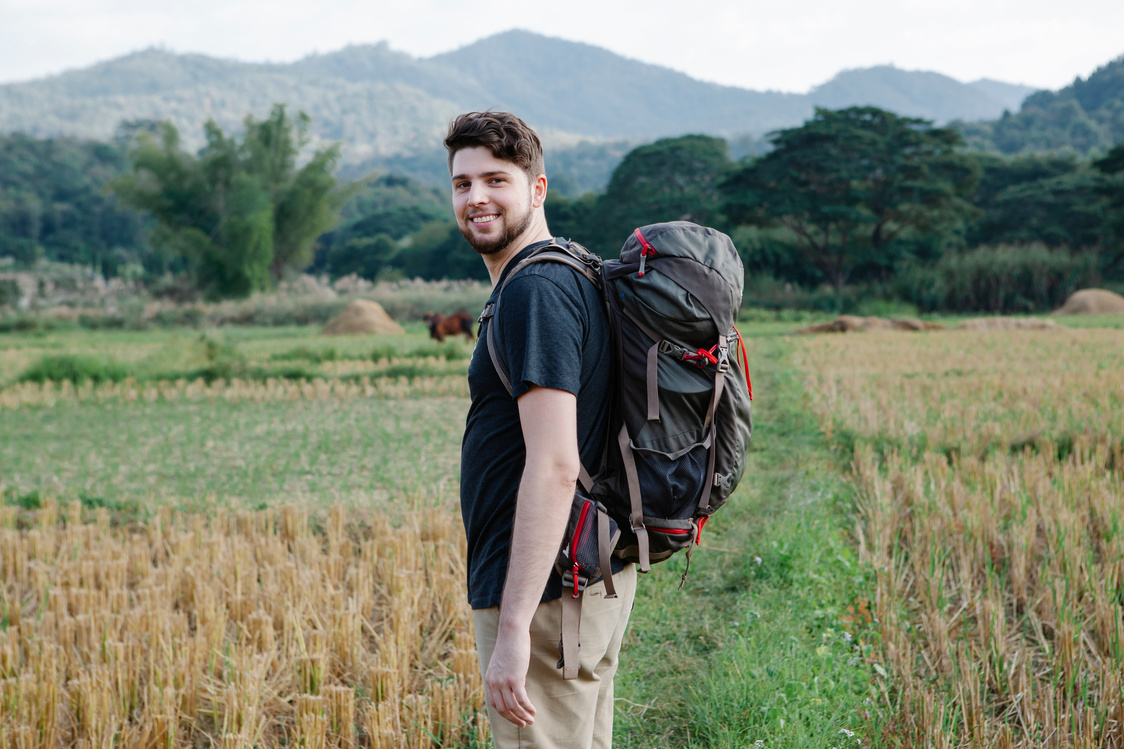 Smiling man with hiking backpack standing in field in countryside in daytime